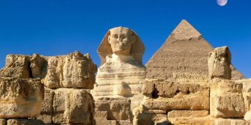 The Great Sphinx, a pyramid and other large stones, in a light colour. the sky is blue and the moon is visible.