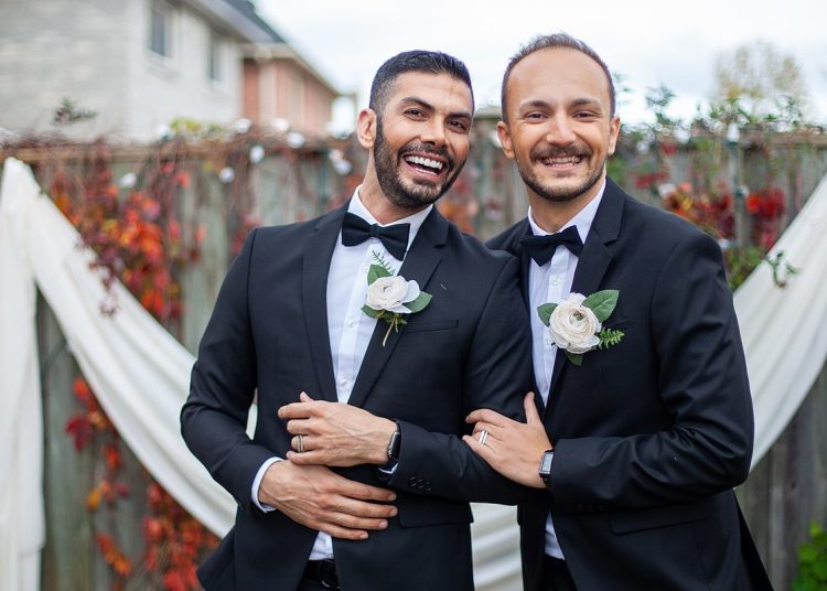 Two men in wedding suits, in front of a white drape. Both wear a white flower in their lapel and look happy