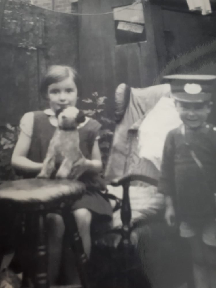 Black-white photo of a young boy wearing jacket and cap, standing next to an older girl who is sitting on a chair and holds a toy dog in her hands