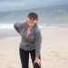 fibromyalgia - woman in grey zip up jacket and black trousers standing on beach holding back and knee, looking exhausted