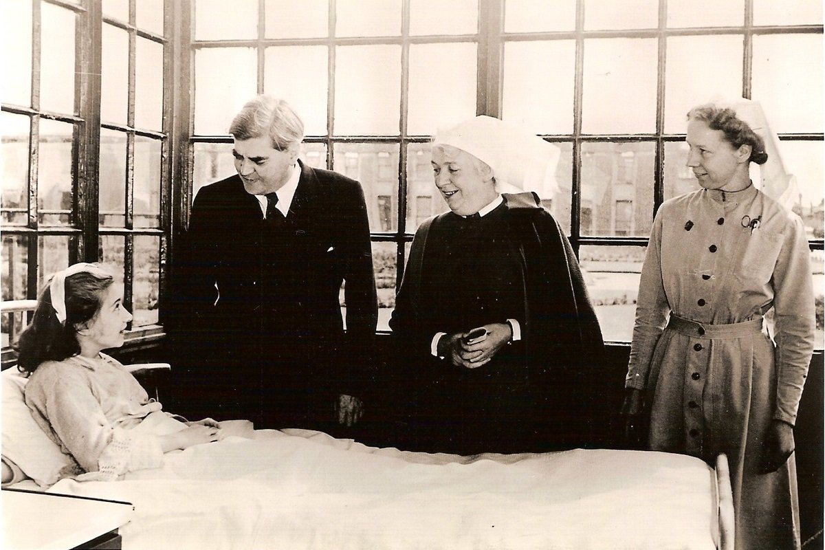 Black and white picture showing a young woman lahing in a hospitak bed, and a man in a suit and two women in oldfashioned nurses uniforms next to the bed.