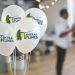 Four white baloons with Central Bylines logo on in the foreground, and some blurred out people in the back of the picture