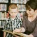 Woman sits next to standing boy and reads to him from a book