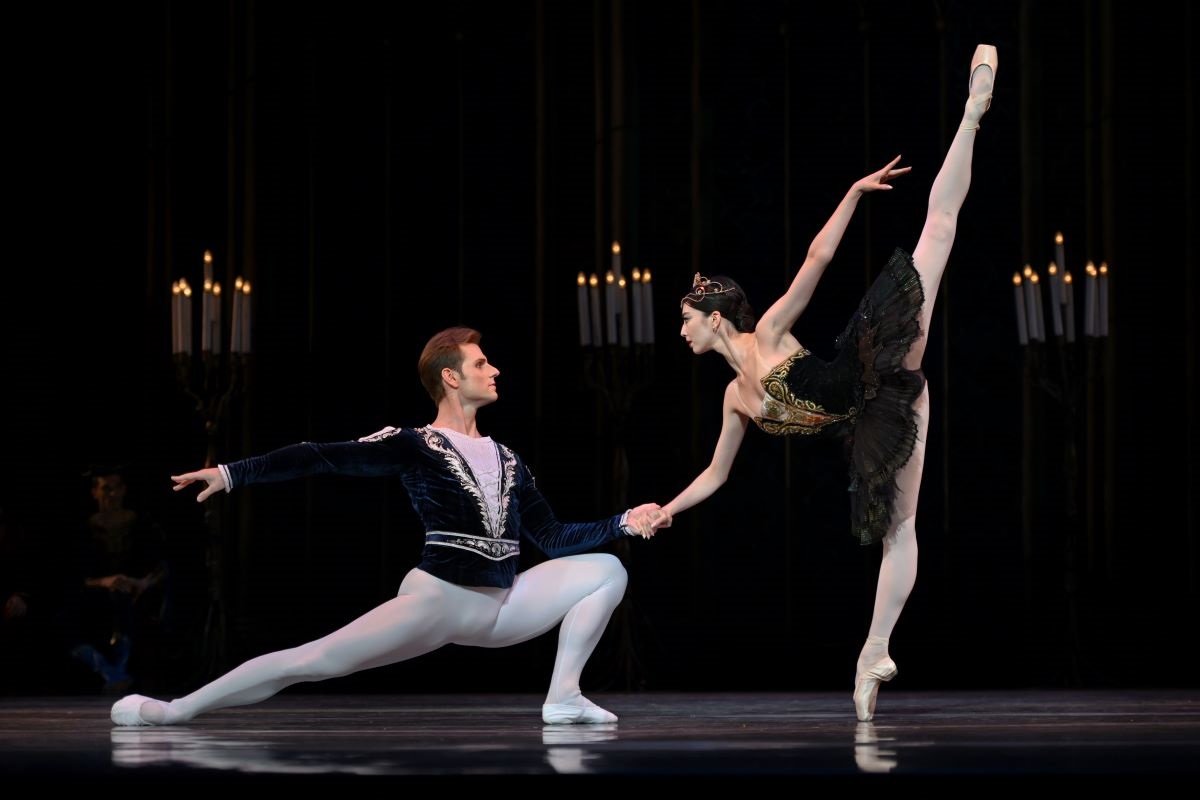 Swan Lake ballet: one dancer doing a split with one leg in the air, and holding the hand of another dancer who kneels