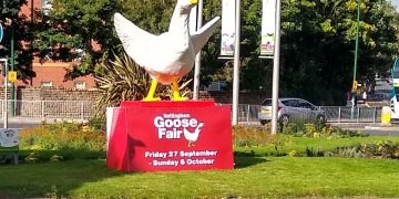 Statue of a white goose with wings spread, standing on a red stand that says 'Goose Fair'. The statue is placed on a roundabout