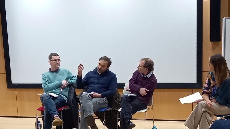Three men sitting on chairs next to each other, the one in the middle is talking and gesturing, and a woman in a chair a bit away from them.