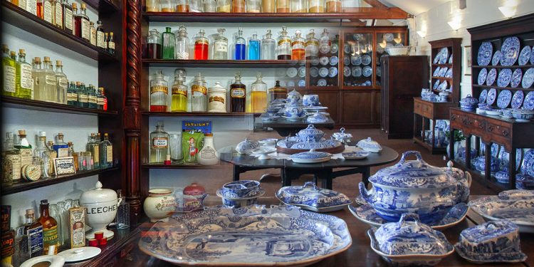 Two image composite split diagonally - top left half is shelves bearing labelled medicine bottles. Bottom right half are table and dressers bearing blue enamelled tableware.