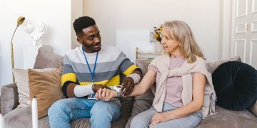 darker skinned young man sits on the couch next to elderly white woman. The man checks the womans blood pressure. both are smiling