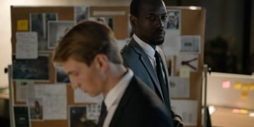 Two detectives stand in front of a corkboard filled with evidence in a dimly lit police station.